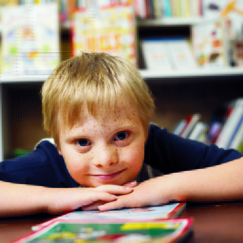 Photo of boy leaning his chin on his hands while sitting at a library table with books behind him