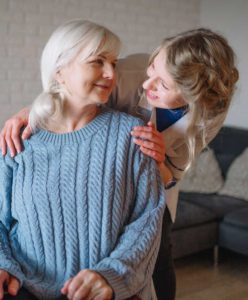 Cargiver leaning over elderly woman with hands on her shoulders.