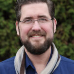 Alan in a blue shirt with a grey and brown scarf in front of trees