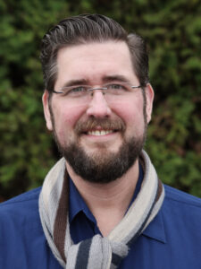 Alan in a blue shirt with a grey and brown scarf in front of trees