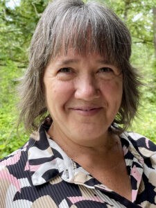 Diane Pearson wearing a white, cream, black, and pink patterned shirt in front of trees