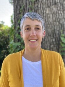 Em Braman in a yellow cardigan and white shirt in front of a tree