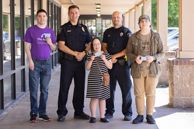 A group of three people with disabilities showing their blue CommCards, standing next to two police officers