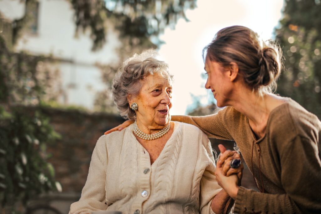 A joyful elderly woman with curly gray hair sits outdoors, wearing a cream cardigan and pearl necklace. She shares a warm moment with a younger woman, who smiles and gently holds her hand, surrounded by greenery and soft sunlight.