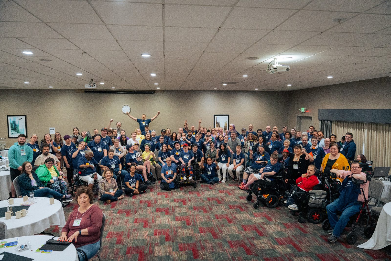A large group of people at the 2025 Oregon Self-Advocacy Convention gathered for a photo in a hotel meeting room. Many wear matching navy blue Disability Rights Are Human Rights shirts. People are standing, seated in chairs, sitting on the floor, and using wheelchairs and mobility devices, smiling and waving at the camera.